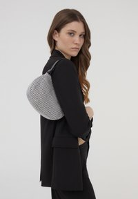 Borsa a tracolla in bianco e nero con texture, forma arrotondata e design a rete, dotata di una tracolla nera e indossata sulla spalla, su uno sfondo neutro.
