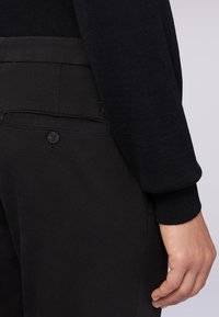 Černé kalhoty s hladkým povrchem a kapsou vzadu s knoflíkem. Přiléhavý černý svetr s žebrovanými manžetami je částečně viditelný.