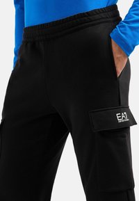 Schwarze Jogginghose aus weichem Stoff, mit elastischem Bund, Seitentaschen und einer aufgesetzten Tasche mit "EA"-Logo an einem Bein.
