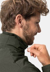 Veste verte avec un col haut, dotée d'une fermeture zippée et d'une doublure en mesh au niveau du col. Le tissu semble lisse et léger.