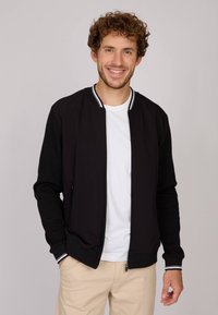 Veste bomber noire en tissu doux avec un col rayé blanc et gris. Présente des poches zippées et des poignets côtelés. Portée sur une chemise blanche.