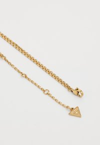 Gouden ketting met schakels, een karabijnsluiting en een driehoekige hanger met de merknaam in reliëf.