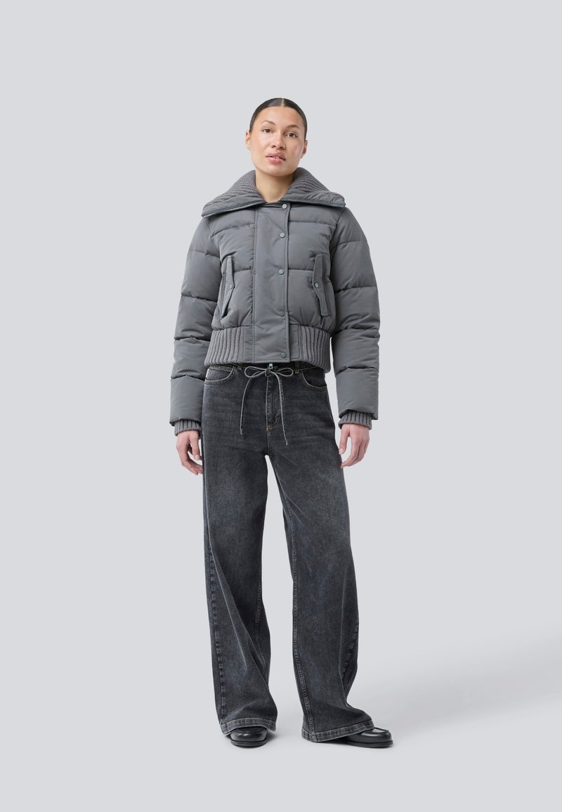 Woman standing wearing a gray puffer jacket with knit cuffs and collar, wide-leg black jeans, and black shoes on a plain gray background.