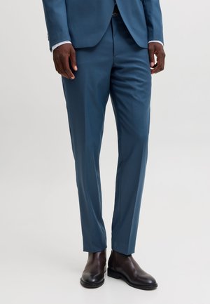 Homme portant un pantalon de costume bleu ajusté avec une veste assortie et des chaussures habillées en cuir marron foncé, se tenant devant un fond uni.