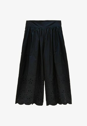 Femme portant un pantalon large court bleu marine avec broderie florale ajourée et des sandales marron à talons avec boucles.
