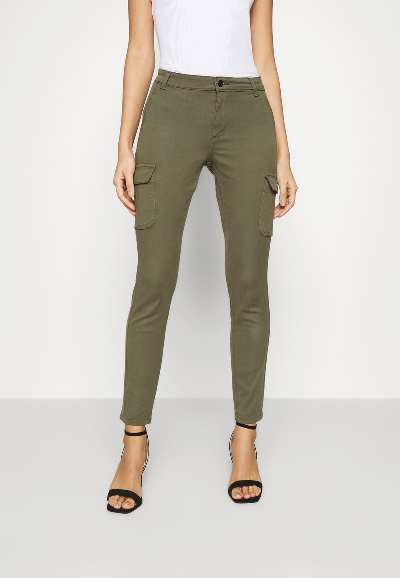 Femme portant un pantalon cargo slim vert olive avec poches à rabat, un haut sans manches blanc et des talons noirs à bride à la cheville, debout devant un fond blanc.