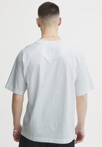 Homme aux cheveux courts et aux oreilles percees, portant une chemise blanche à manches courtes vue de dos sur fond blanc.