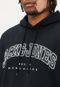 Svart huvtröja med dragsko, med stor vit text som läser "JACK&JONES EST. 10 WORLDWIDE" i fet stil. Mjuk material.