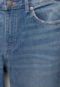 Jean en denim bleu en gros plan montrant la poche avant, les détails des coutures et le bouton en métal avec gravure de la marque.