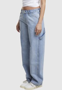 Personne portant un jean ample bleu clair avec poche latérale, des baskets blanches et un top court blanc, debout devant un fond uni.
