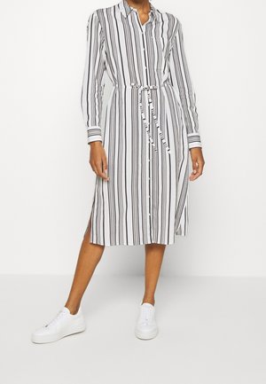Femme portant une robe-chemise blanche à rayures verticales noires, fermeture par boutons, ceinture à la taille, fentes latérales, et baskets blanches.