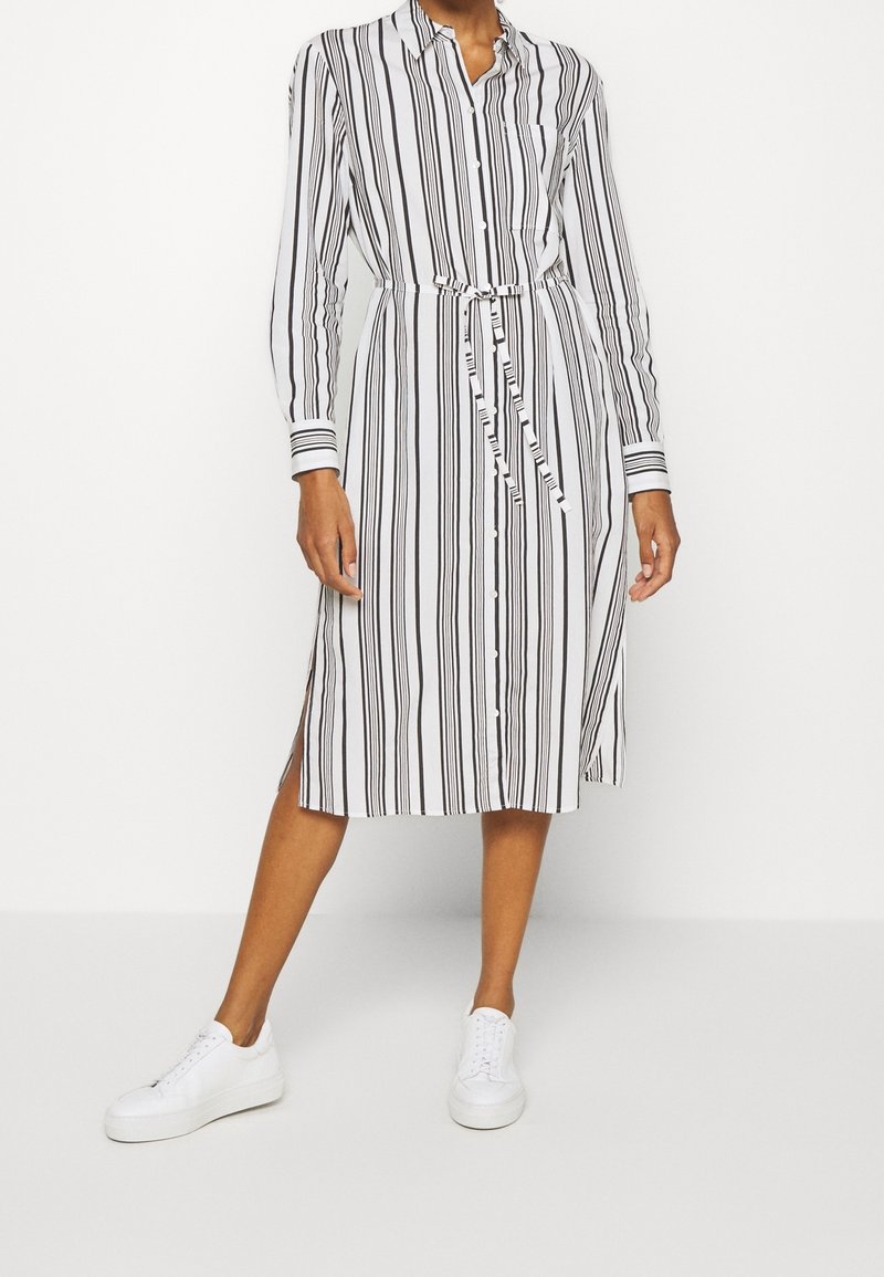Femme portant une robe-chemise blanche à rayures verticales noires, fermeture par boutons, ceinture à la taille, fentes latérales, et baskets blanches.