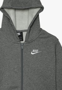 Grå hoodie i strukturerad tyg, med hel dragkedja, huva och en vit broderad Nike-logotyp på vänster bröst.
