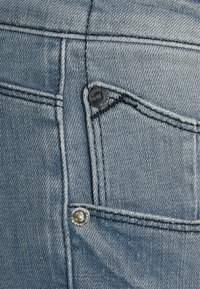 Ljusblå denimtyg med en liten framficka, silvertråd och metallhardware, inklusive en märkesknapp och nitat detalj.