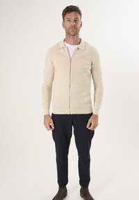Beige ritscardigan met een kraag, ribbelstructuur, getailleerde pasvorm, gecombineerd met marineblauwe broek en bruine laarzen.