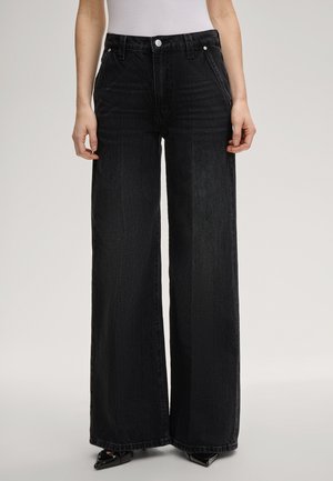 Kobieta nosząca czarne, luźne jeansy z wysokim stanem, przednimi kieszeniami, srebrnym guzikiem i szerokimi nogawkami, w połączeniu z czarnymi, spiczastymi butami.