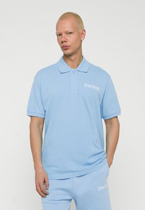 Jeune personne aux cheveux blonds courts portant un polo bleu clair Bruno Banani et un pantalon assorti avec un texte de logo blanc.