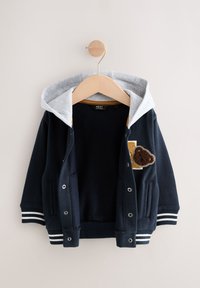 Giacca con cappuccio blu navy con fodera grigia, polsini a righe bianche e blu navy e toppa con orso sul petto. Chiusura con bottoni a scatto.