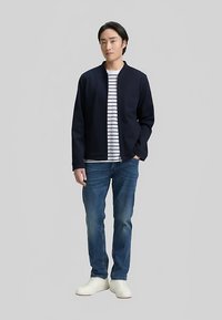 Marineblauw jasje over een wit en marineblauw gestreept shirt, gecombineerd met blauwe jeans en witte sneakers; casual ontwerp met een ontspannen pasvorm.