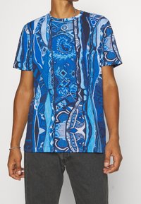 Man med kortärmad blå mönstrad skjorta med abstrakta vertikala och geometriska mönster, kombinerad med mörkgrå byxor.