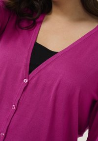 Fuchsia cardigan med V-hals och fem knappar framtill. Stickad textur, lätt tyg, bärs över en svart topp.