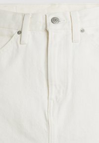 Gros plan sur le devant d’un jean en denim blanc montrant un bouton, une fermeture éclair, des passants de ceinture et une partie des poches avant avec des rivets métalliques.
