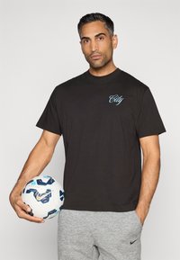 Čierne bavlnené tričko s svetlomodrým logom "City" na hrudi, v kombinácii so sivými teplákmi. Osoba drží vzorovanú futbalovú loptu.