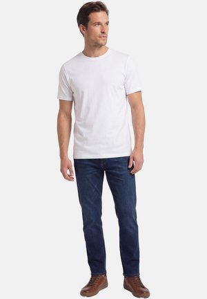 Man standing, wearing plain white short-sleeve t-shirt, dark blue jeans, and brown leather sneakers, looking slightly right.