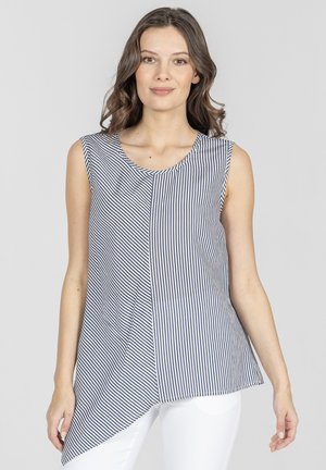Kobieta ubrana w bezrękawnikową, asymetryczną bluzkę w pionowe paski w kolorze granatowo-białym oraz białe spodnie, stojąca na jednolitym tle.