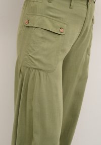 Olivově zelené cargo kalhoty s boční kapsou a dřevěnými knoflíkovými detaily a nařasenými švy blízko lemu. Měkká, lehká tkanina.