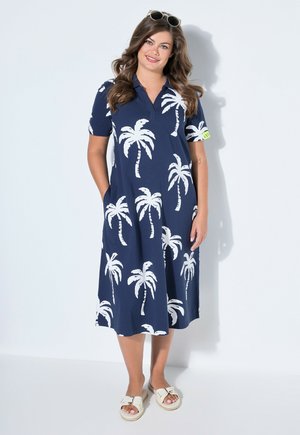 Donna con un vestito midi blu navy con stampe bianche di palme, occhiali da sole sulla testa e sandali slip-on bianchi, in piedi davanti a uno sfondo neutro.