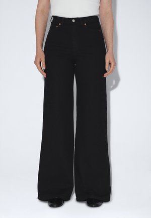 Kvinde iført sorte viddebenbukser med højtaljet snit og kobberfarvede nitter, kombineret med en hvid ærmeløs top og sorte sko, stående mod en hvid baggrund.