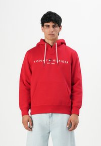 Un uomo che indossa una felpa rossa di Tommy Hilfiger con cordini bianchi e jeans azzurri chiari, in piedi davanti a uno sfondo bianco uniforme.