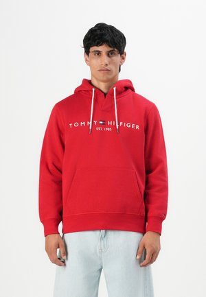 Muškarac u crvenoj Tommy Hilfiger dukserici s bijelim konopcem i svijetloplavim farmerkama, stoji naslonjen na običnu bijelu pozadinu.