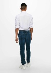 Camisa blanca de hombre con botones combinada con jeans oscuros de corte slim y zapatillas blancas, vista por la parte de atrás, mostrando un diseño sencillo.