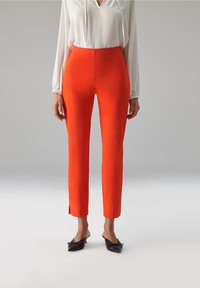 Feloranje nauwsluitende broek met een hoge taille, zijslitsen aan de zoom, gecombineerd met een witte blouse en zwarte instapschoenen. Soepele textuur.