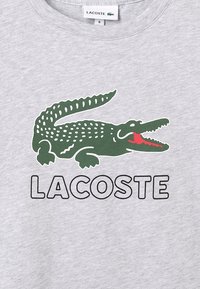 Harmaa collegepaita, jossa on vihreä krokotiililogo punaisin yksityiskohdin ja musta teksti "LACOSTE" alla. Pehmeä pinta, rento muotoilu.