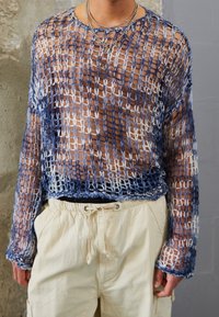 Personne portant un pull en maille ouverte bleu et blanc sur peau nue, associé à un pantalon beige clair à cordon de serrage et des colliers en chaînes d'argent superposés.