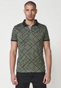 Olivinės spalvos polo marškiniai su juodu geometriniu raštu, trumpomis rankovėmis ir juodu apykakle. Yra trijų mygtukų užsegimas ir mažas logotipo detalė.