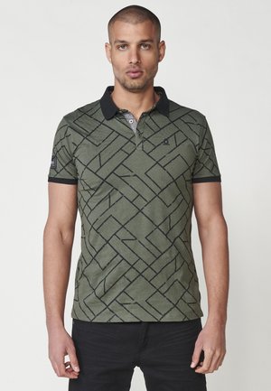 Camisa polo verde oliva con patrón geométrico negro, mangas cortas y un cuello negro. Cuenta con una tapeta de tres botones y un pequeño detalle de logo.