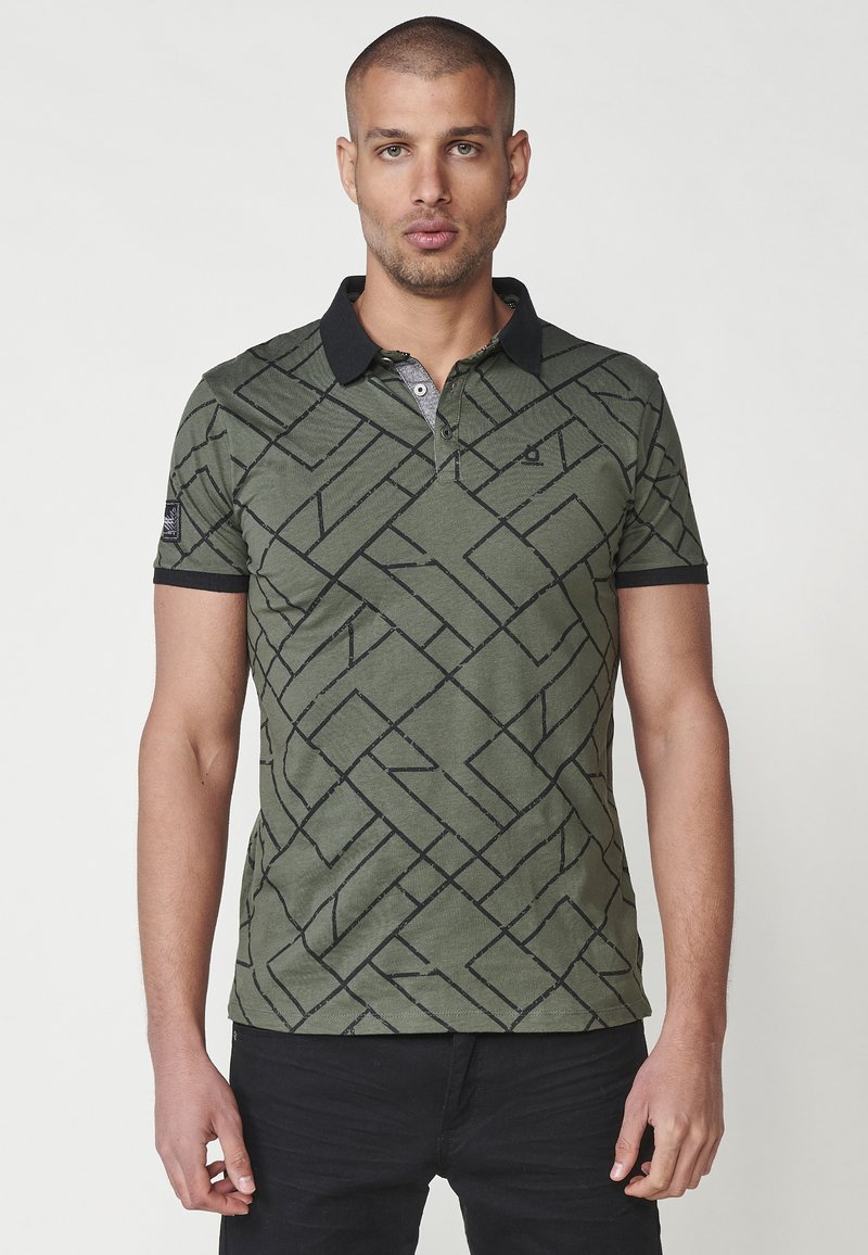 Olivinės spalvos polo marškiniai su juodu geometriniu raštu, trumpomis rankovėmis ir juodu apykakle. Yra trijų mygtukų užsegimas ir mažas logotipo detalė.