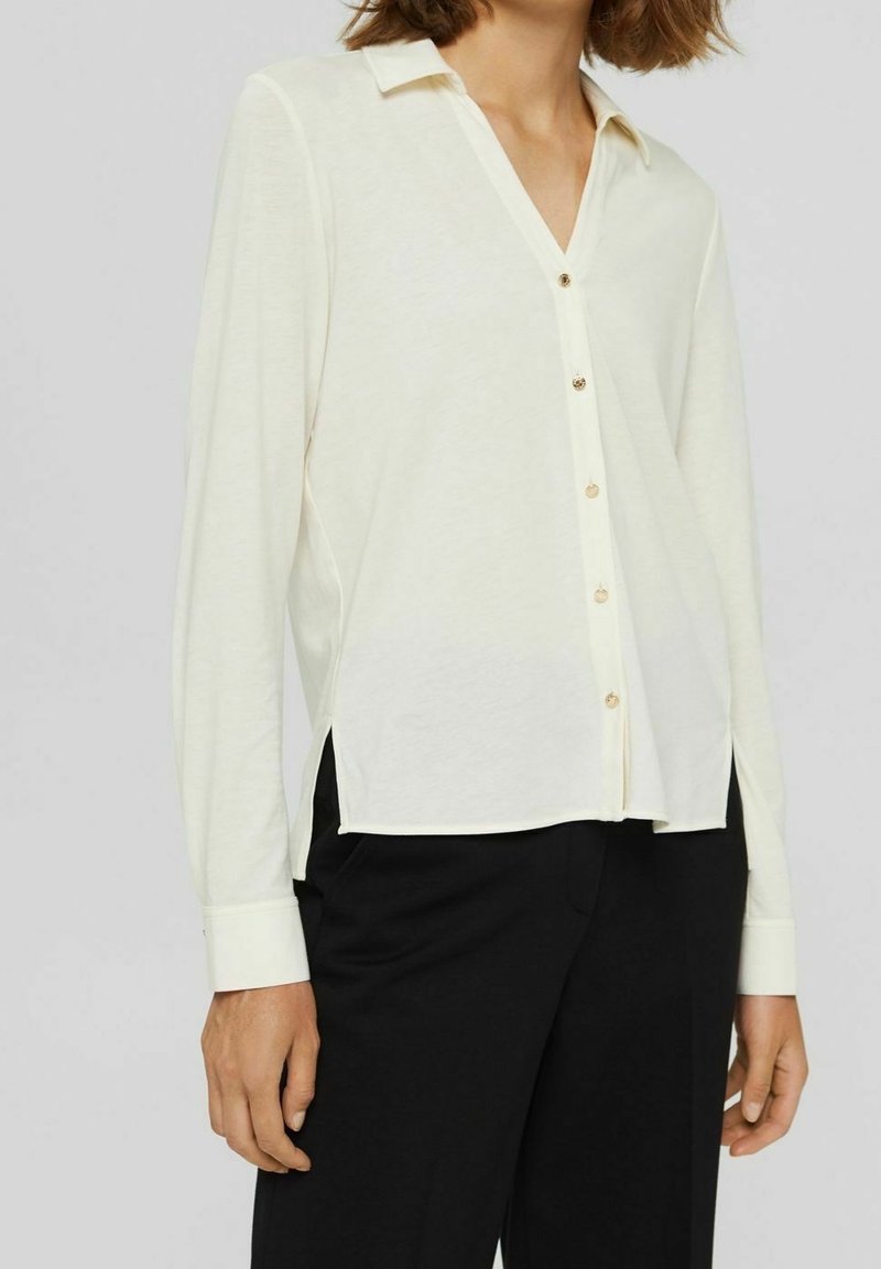 Femme portant une chemise blanche à manches longues boutonnée avec col et un pantalon noir, debout devant un fond uni.