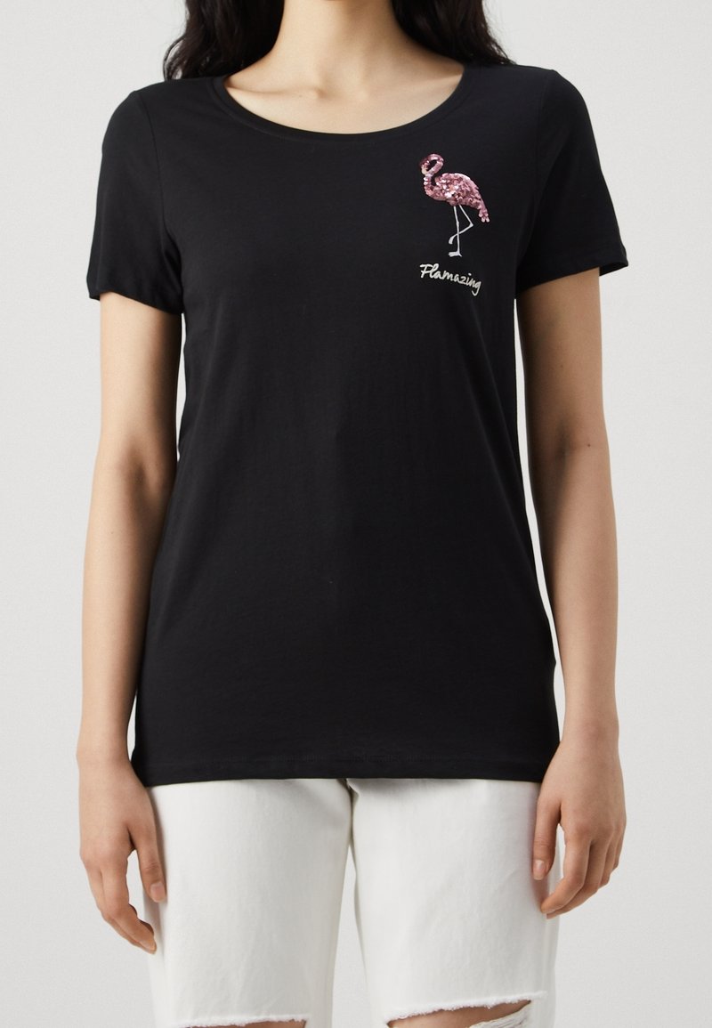 Svart kortärmad t-shirt med rosa paljetflamingo och texten "Flamazing" som bärs med vita rivna jeans på en stående person.