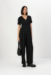 Combinaison noire à manches courtes, décolleté croisé, pantalons à larges jambes, associée à des chaussures à plateforme noires épaisses et un petit sac à main noir.