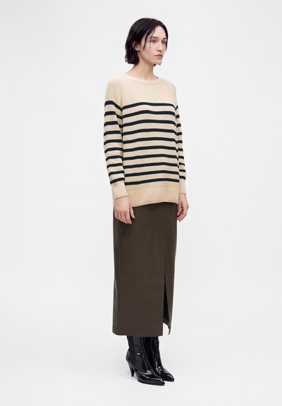 Femme portant un pull beige à rayures horizontales noires, une longue jupe marron foncé avec une fente à l'avant, et des bottines noires à talons.