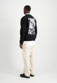 Felpa nera con stampa grafica sul retro, abbinata a pantaloni cargo beige larghi e scarpe nere. Tessuto morbido, design casual.
