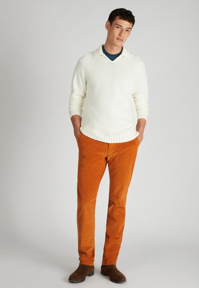 Weißer Strickpullover mit geripptem Saum, orangefarbene Cordhosen und braune Wildleder-Stiefeletten. Lässiger Schnitt mit subtilen Texturkontrasten.