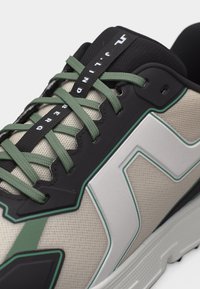 Béžová teniska so zelenými šnúrkami, čiernymi očkami a bielym geometrickým bočným dizajnom, doplnená čiernou páskou na jazyku s logom značky.