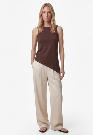 Vrouw met blond haar die een mouwloos bruin asymmetrisch topje, brede beige broek en zwarte sandalen draagt, staat recht naar voren.