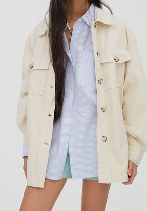 Femme portant une veste beige oversize par-dessus une chemise rayée bleue et blanche avec un short vert clair, debout devant un fond uni.